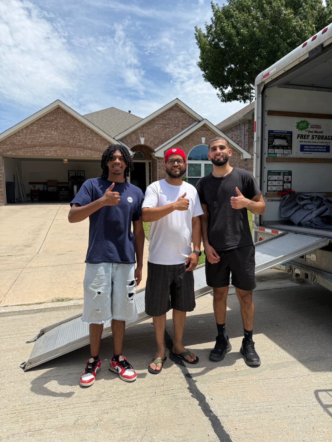 Two Nahas Moving movers giving a thumbs up with a customer in a driveway beside an open U-Haul truck and loading ramp